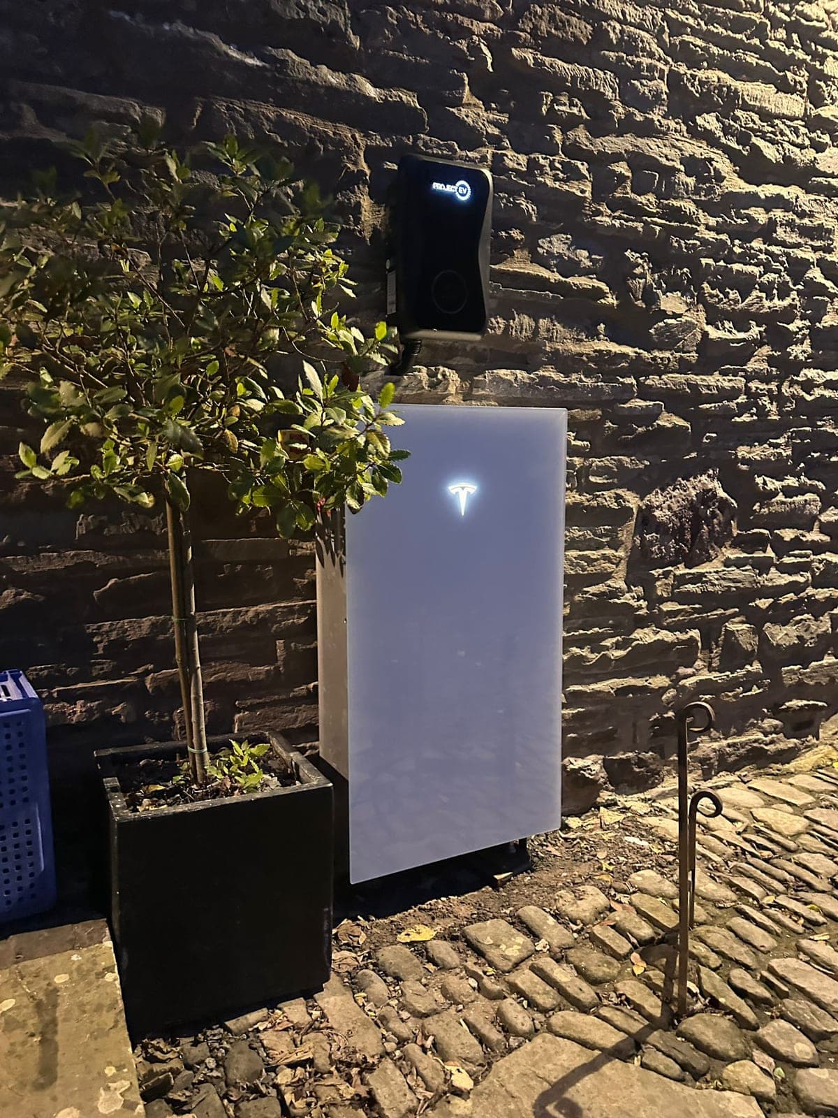Tesla Powerwall installed outside at night, T logo lit, alongside the front door planter